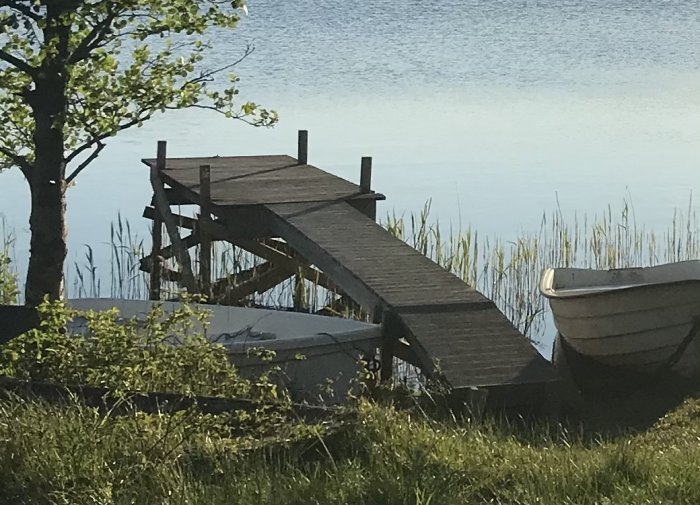 Reparerad träbrygga vid sjö med ny stötta och reglar, bredvid roddbåt och grönska.