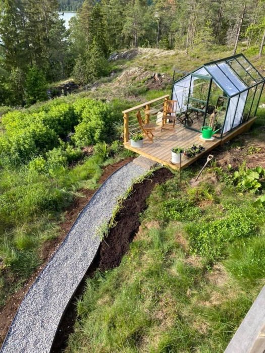 Ny grusgång leder till växthus med trädäck i en trädgård med utsikt över sjö.