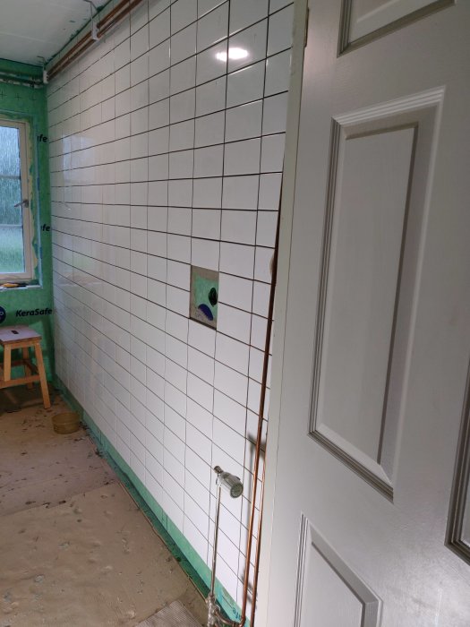 Nästan färdiglagda vita kakelplattor på vägg med synlig rörgenomföring och utanpåliggande rör.