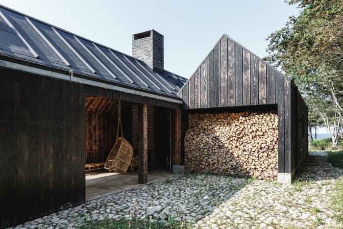 Trähus med listtäckt papptak och vedstapel vid fasaden, designat av Lendager Group.