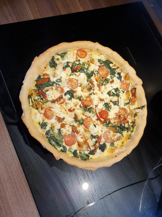 Hembakad kyckling- och spenatpaj med fetaost och tomater på en mörk yta.