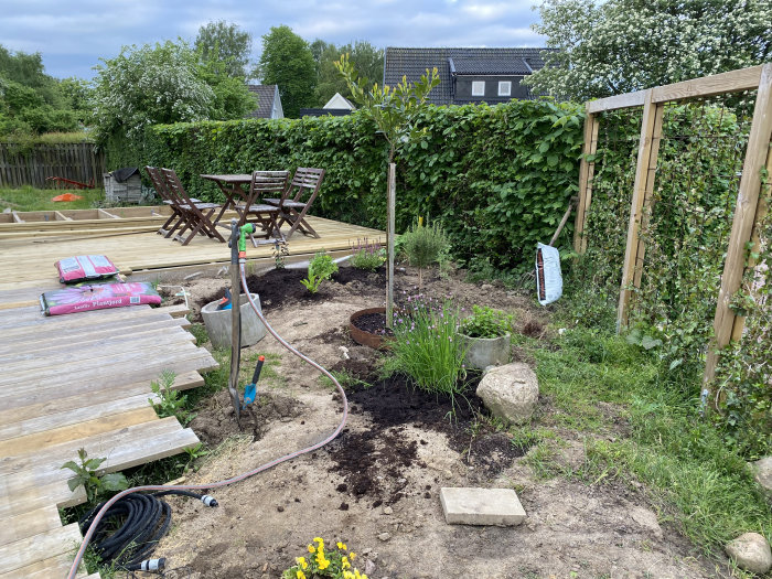 En trädgård under anläggning med jordytor förberedda för plantering av kryddor och blommor, intill en träaltan och trädgårdsmöbler.