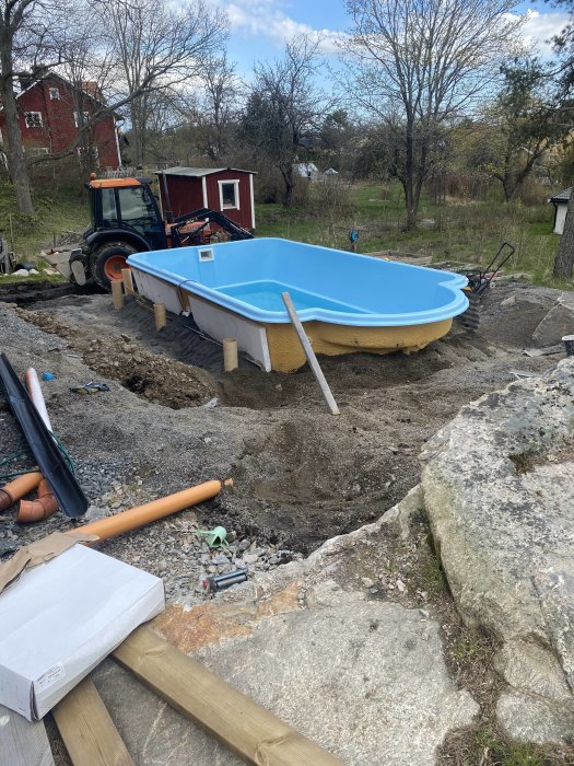 Blå pool under installation i utegrävd trädgård med motfyllning och rörarbete synligt, samt traktor i bakgrunden.