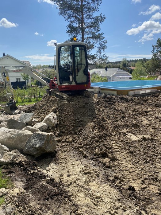 Minigrävare vid pågående byggprojekt för altan nära en halvmonterad pool, omgiven av sten och jord.