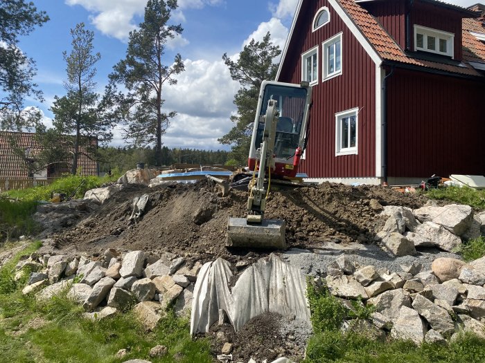 Minigrävare vid pågående markarbete nära röd stuga med pool i bakgrunden, motfyllning av altan grund.