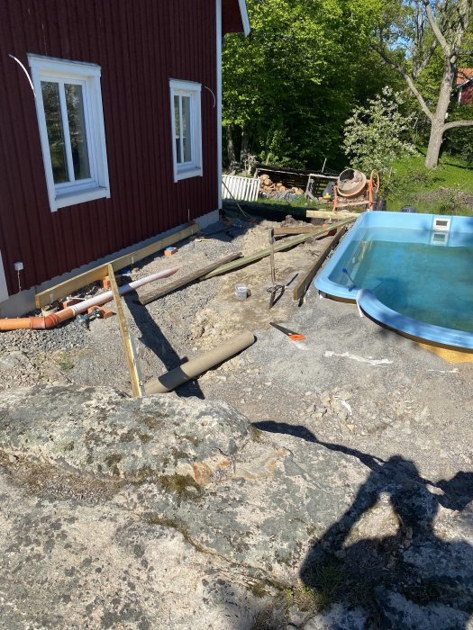 Byggplats med pågående arbete vid en pool och husvägg, med isolerade rör och stommen till en altan under konstruktion.