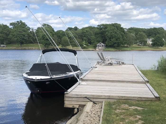 Motorbåt förtöjd bredvid en träbrygga vid en lugn sjö med grönskande träd i bakgrunden.