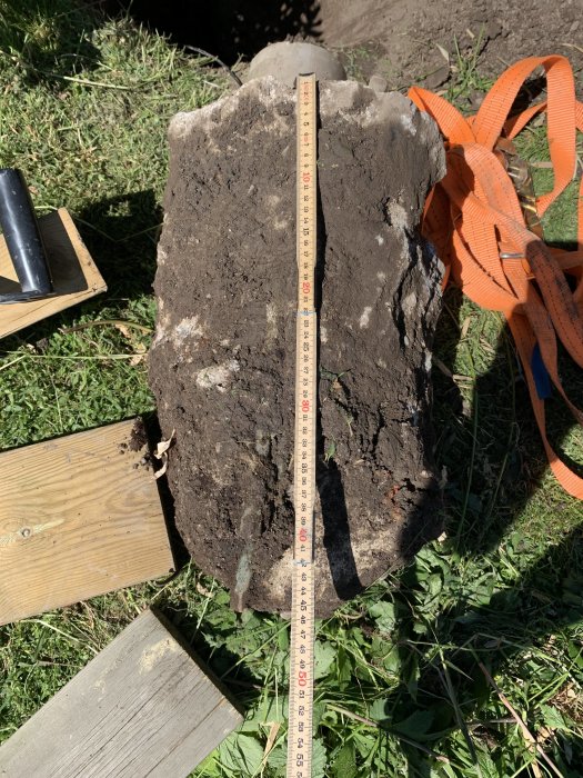 Måttstock står mot en stor sten i gräset som mäter nästan 50 cm i höjd, med byggmaterial bredvid.