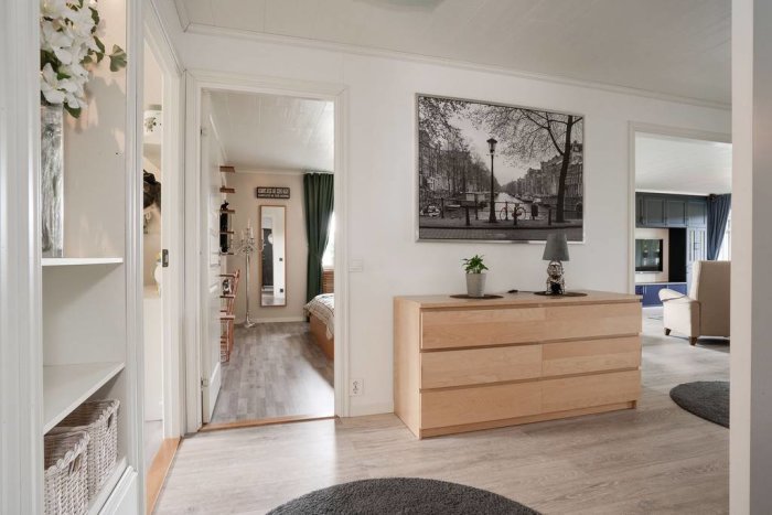 Interiörvy av ett modernt vardagsrum med träbyrå, svartvitt konstverk och öppen dörr till sovrum.