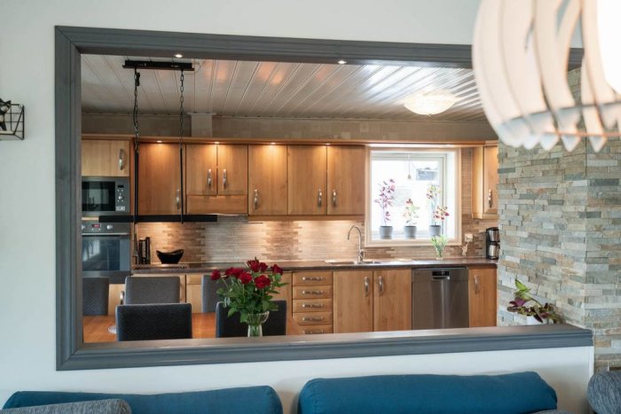Inblick i ett modernt kök med träskåp och rostfria vitvaror genom en spegelvägg.