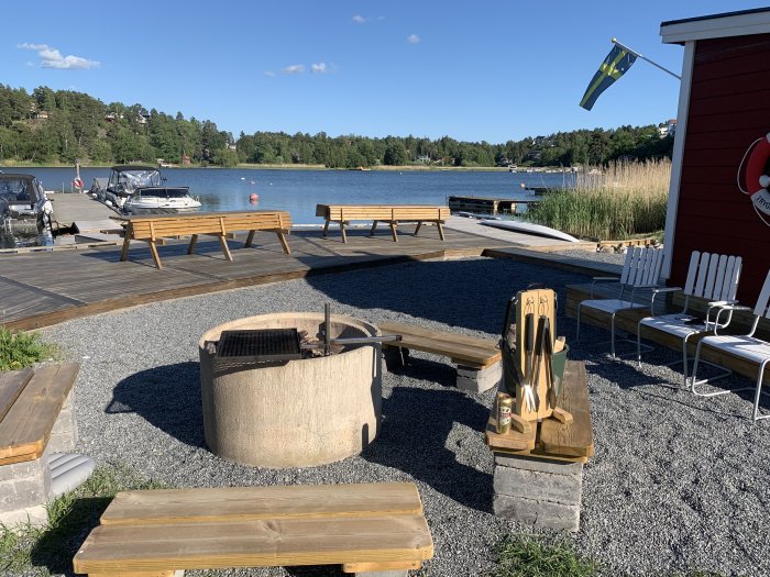 Sommardag vid sjön med brygga, badtunna, grillplats, stolar och en svensk flagga i bakgrunden.