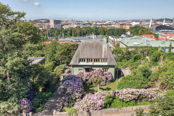 Flygbild över ett traditionellt hus omgivet av grönska och blommande buskar med Göteborg i bakgrunden.