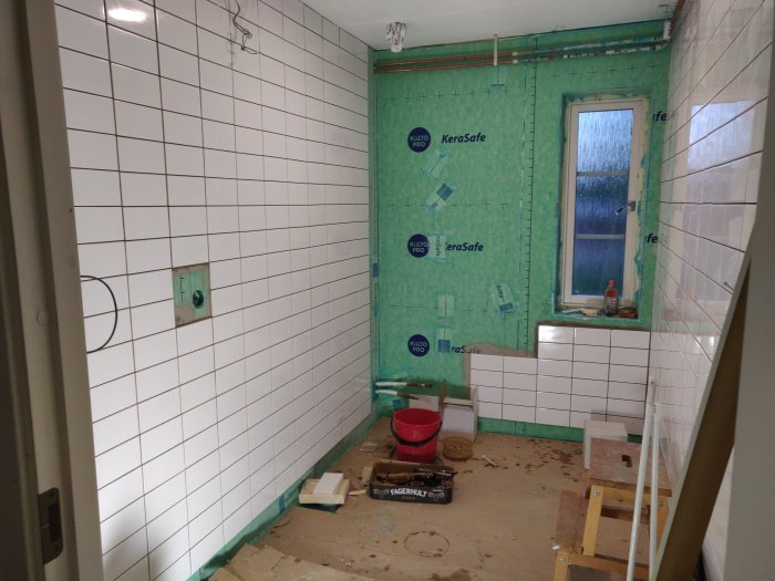 Delvis kaklad vägg i badrum med små vita plattor, oavslutade hörn och fönster med isoleringsmaterial.