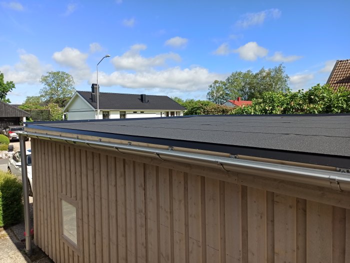 Nytt tak på garage med tvålagstäckning och hängrännor, i bakgrunden syns hus och grönska under en delvis molnig himmel.