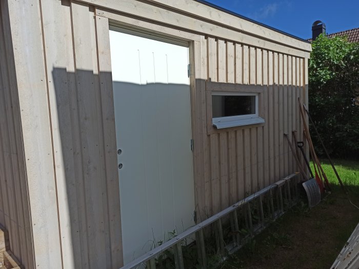 Ny stående träpanel på garagevägg med vit garageport och litet fönster, omgiven av byggmaterial.