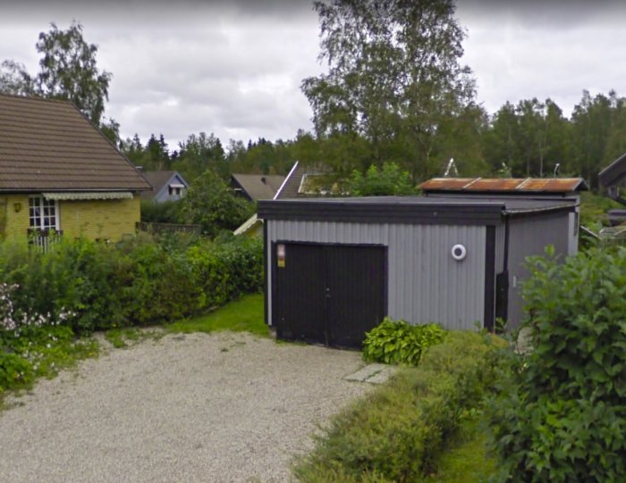 Ett grått garage med platt tak och vita detaljer före ombyggnation, intill en uppfart och grönska.