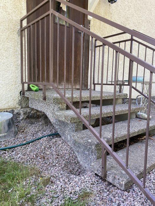 Udendørs betontrappe med brun gelænder ved en indgangsdør. Trappen er på et underlag af grus og græs.