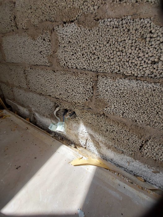 Delvis borttaget golv i källare med synliga vattenskador och blött material mot en lecablocksvägg och en lös elkabel.