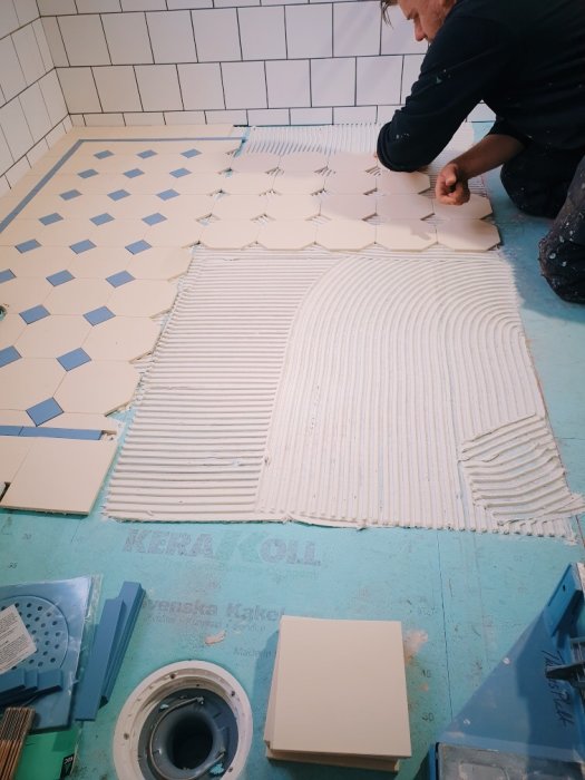 Person som installerar kvadratiska kakelplattor på golv med mörtel, omgiven av byggmaterial.