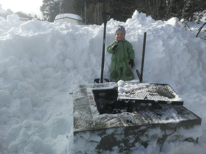 Ett barn står bredvid en ca 55 cm hög eldstad omgiven av en stor snöhög.