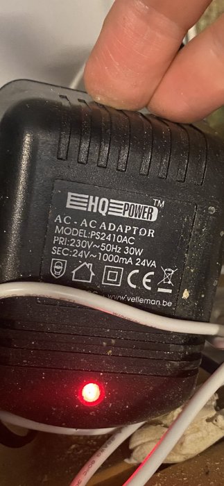 Hand som håller en inkopplad AC-adapter med specifikationsetikett och tänd röd indikatorlampa.