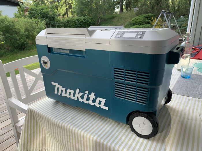 En Makita-kylbox på ett randigt bord ute på en veranda med gräsmatta i bakgrunden.