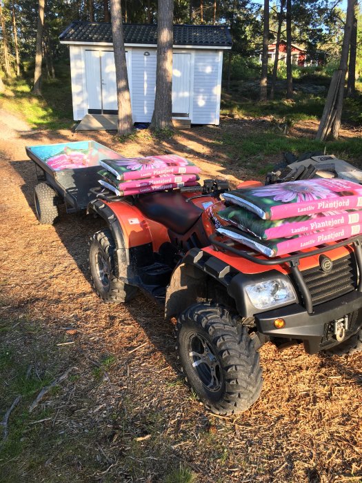 ATV med släpkärra överlastad med rosa säckar med planterjord framför en vit skjul i en trädgård.