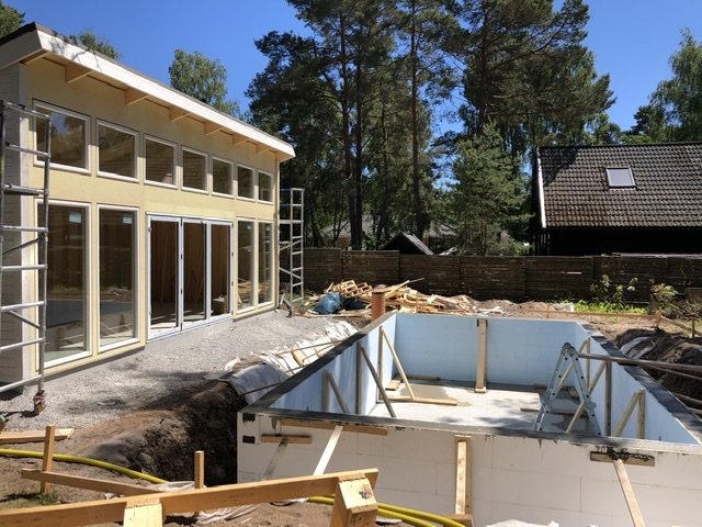 Delvis färdigt hus med vikdörrar och en obyggt poolområde i förgrunden under en klar himmel.