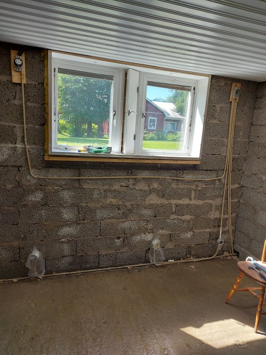 Nyrenoverat källarrum med upprivet golv, exponerad betongvägg, ett fönster och elektriskt kabel.