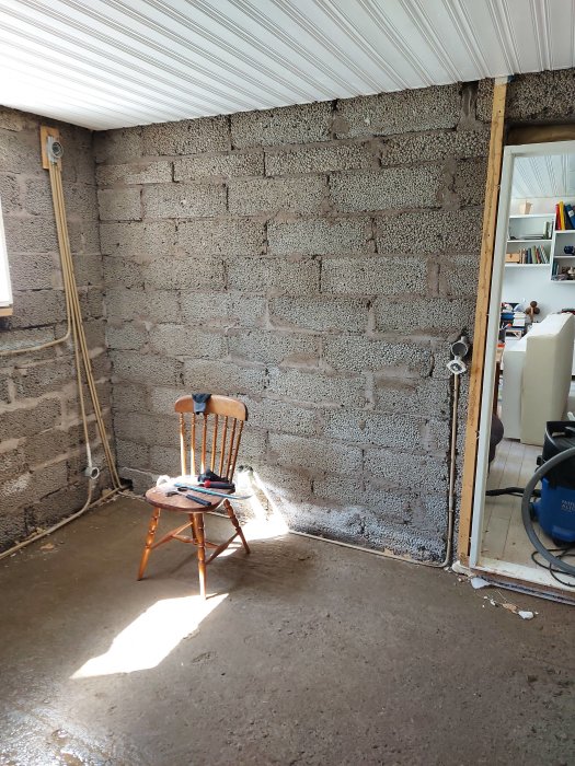 Ett upprivet golv i en källare med en trästol, elledningar längs en blockvägg och renoveringsverktyg.