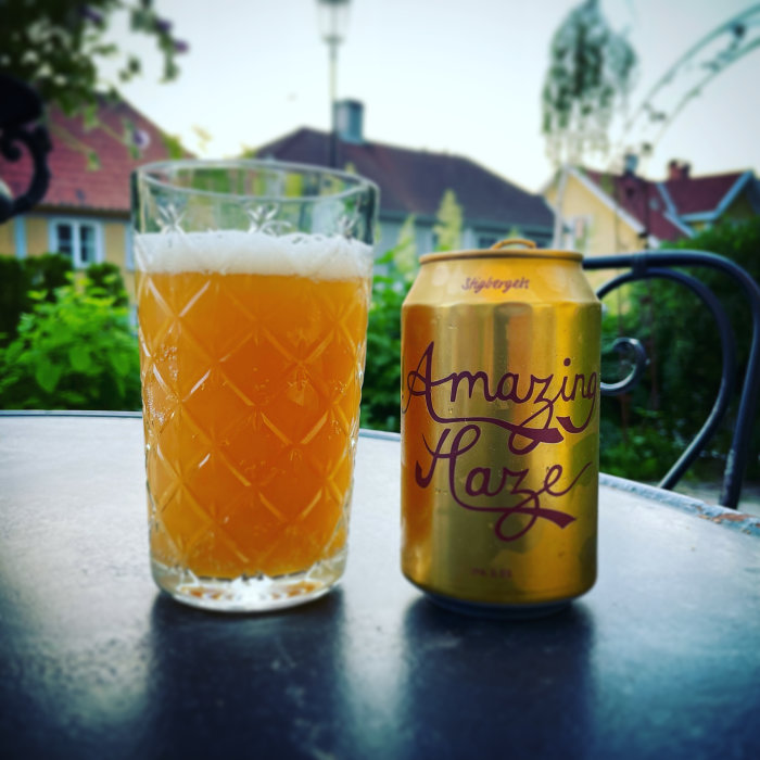 Ölglas med skum och en burk märkt "Amazing Haze" på ett utomhusbord, bokeheffekt i bakgrunden.