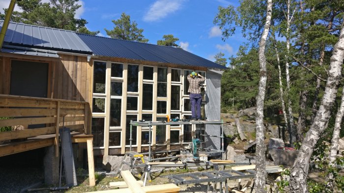 Renovering av växthus med person som fäster fönster på en trästomme, verktyg och trämaterial syns framför.