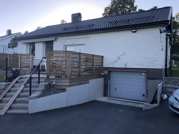 Husets fasad med synliga fogarbeten, trädäck med staket och en garageuppfart.