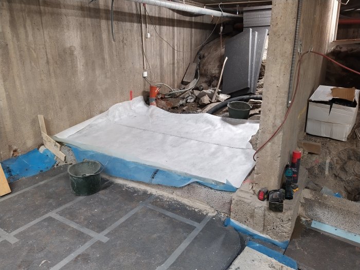 En rörig källare under renovering med skyddsplast på golvet, byggmaterial och verktyg utspridda.