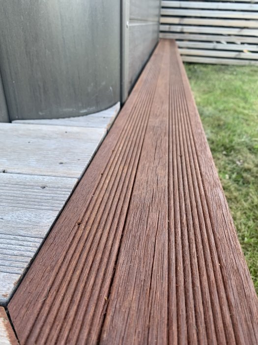 Närbild av en mörkbrun burmateak trallbräda med synlig textur bredvid en grå vägg och gräs i bakgrunden.