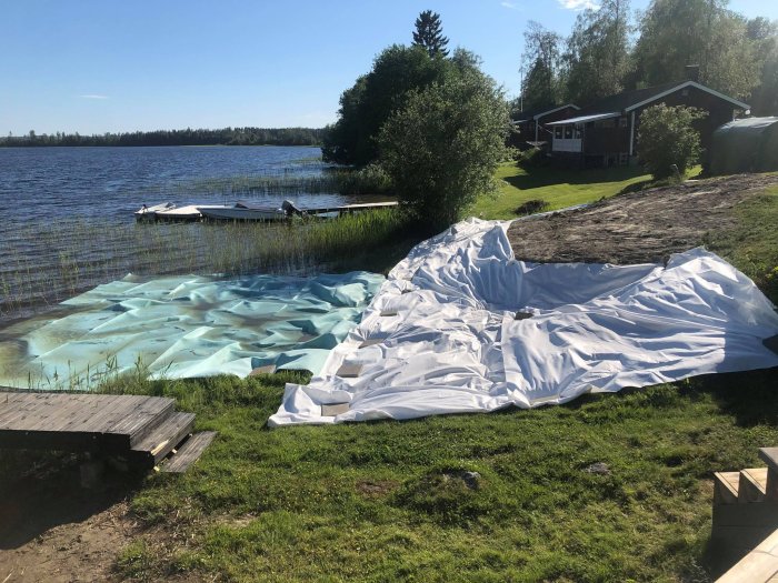 Plastduk utlagd på marken vid sjö för anläggning av sandstrand, med brygga och båt i bakgrunden.