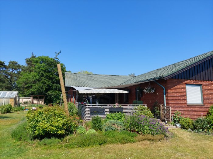 Trädgård med nyuppsatta pelare för solsegel vid en röd tegelvilla under en klarblå himmel.