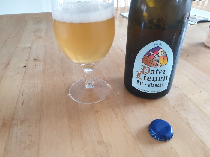 Ett glas witbier på ett bord bredvid en öppnad flaska märkt 'Pater Lieven Wit-Blanche' och en blå kapsyl.