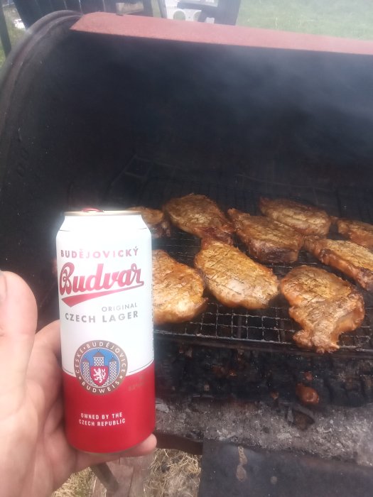 Hand håller en burk Budvar-öl framför grill med stekande köttstycken.