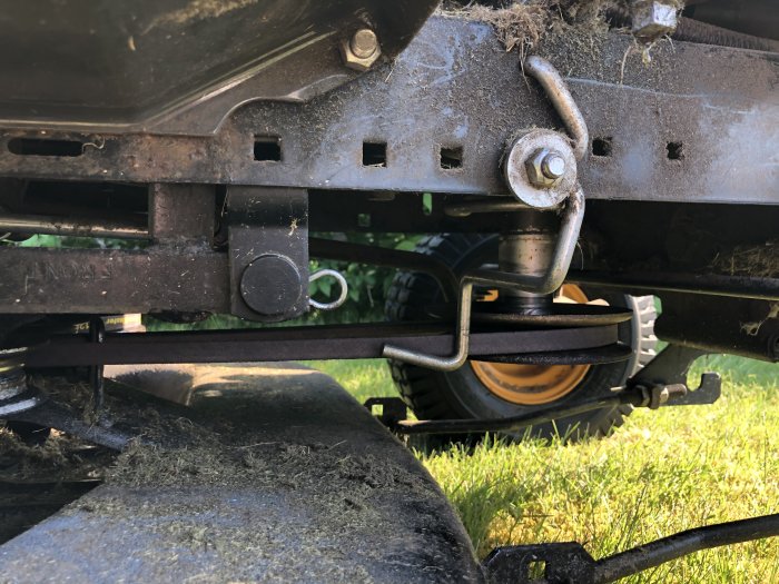 Undersida av en gräsklippare som visar drivrem och dess spännmekanism med synliga gräsklippningsrester.