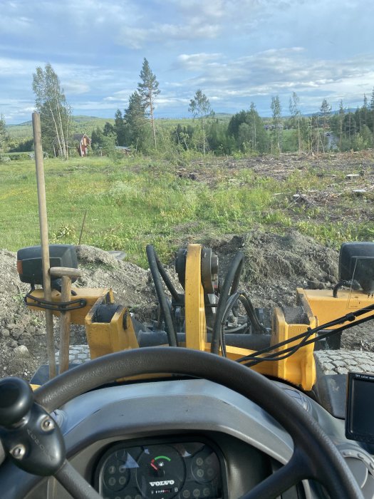 Utsikt från grävmaskinens hytt över område för gräsmattan med skog och hus i bakgrunden.