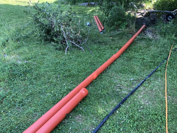 Långa orangea dräneringsrör ligger på en gräsmatta med beskuren vegetation i bakgrunden.