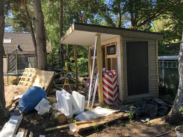 En person bygger ett pumphus i trä för en pool, med byggmaterial och verktyg runtomkring.
