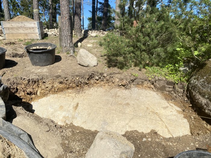 Jordig mark med stor sten och uppgrävd yta för plantering nära barrträd och stenar.