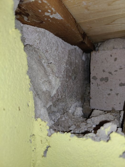 Grå betong och brunt trä syns bakom en gul isoleringsmaterialskikt i en byggkonstruktion.