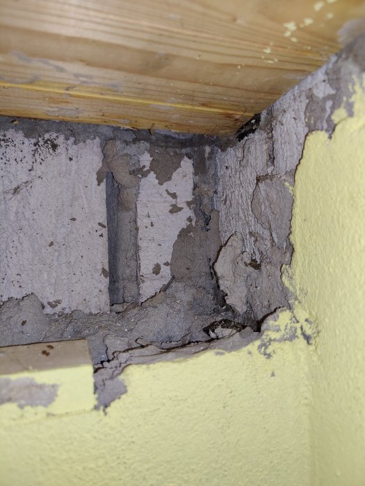 Skadat område med sprickor och löst puts vid träbjälke och betonggolv, behov av renovering.