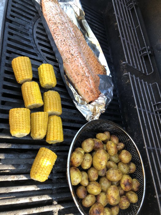 Grillad hel laxfilé i folie, majs på kolvar och stekt potatis på en grill.