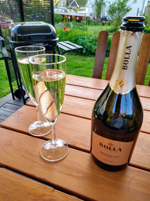 Två champagneflöjtar med bubbel och en flaska Bolla Prosecco på ett träbord utomhus.