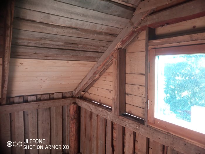 Rivet hörn av trähus med öppna takbjälkar och synliga väggsyllar nära fönster.