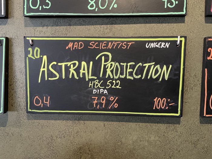 Svart tavla med ölsorten "Astral Projection" skrivet med kritor, inklusive detaljer om alkoholhalt och pris.